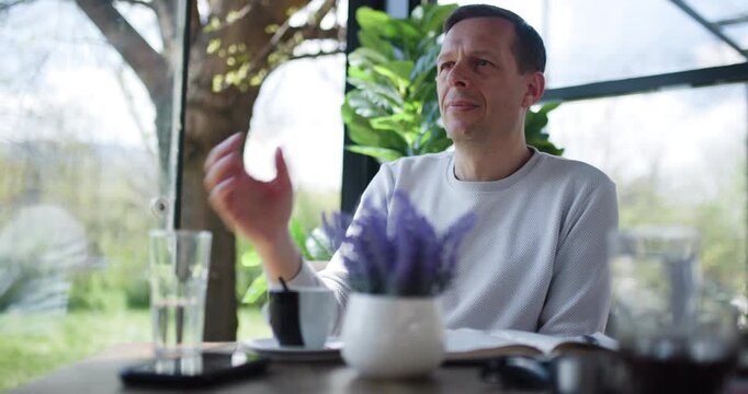 A man sits at a table in a cafe, sipping a beverage while surrounded by plants. The ambiance is peaceful, inviting deep thoughts and relaxation during a bright afternoon