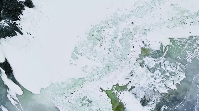Top-down drone view of frozen sea meeting a dark rocky shoreline, with cracked ice sheets and green algae patches forming abstract winter textures and copy space.