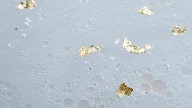 Gold leaf fragments and air bubbles drift through a pale blue cosmetic fluid in a laboratory setting. Close-up view.