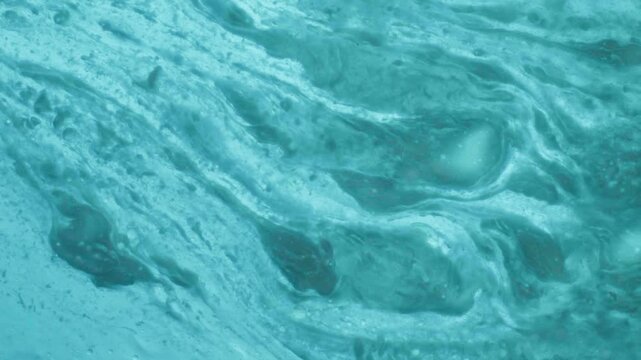 Swirling turquoise and white liquids mixing together to form abstract patterns in a lab setting. Microscopic view of chemical reactions for cosmetic product development.