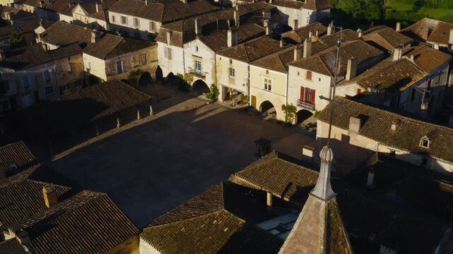 Tiled rooftops of traditional houses surround the central square of Monpazier village in Dordogne. Drone shot moving over a stone spire.