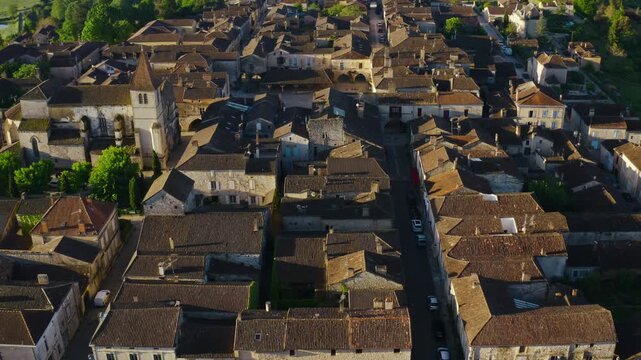 Monpazier bastide in Dordogne featuring historic stone buildings and a church tower. Traditional rooftops define the medieval village landscape. High angle drone shot.