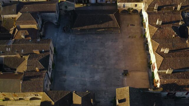 Close view of the ancient stone buildings and arched walkways surrounding the main square of Monpazier. Top-down drone movement.