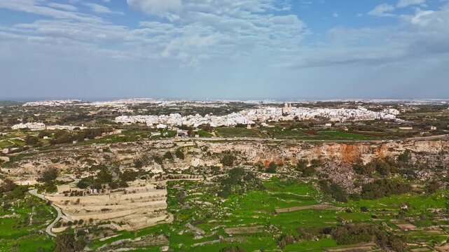 Dingli Cliffs in Malta rise above terraced fields and a rural village with traditional Mediterranean architecture, captured in a panoramic aerial hyperlapse under a partly cloudy winter sky.