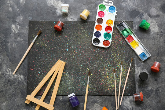 Colorful paint splatter on black paper with brushes, watercolor palette, gouache jars and wooden easel on gray background, top view creative art workspace mockup