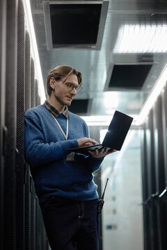 Caucasian middle aged man standing in server room using laptop computer, wearing eyeglasses and identification badge, monitoring equipment and performing technical maintenance tasks, vertical
