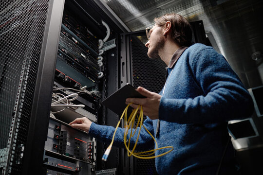 Middle aged Caucasian man working in server room holding tablet and yellow network cables, inspecting server rack equipment, focusing on data center maintenance and technology management
