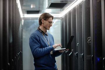 Caucasian middle aged man working on laptop inside server room, standing between rows of server...
