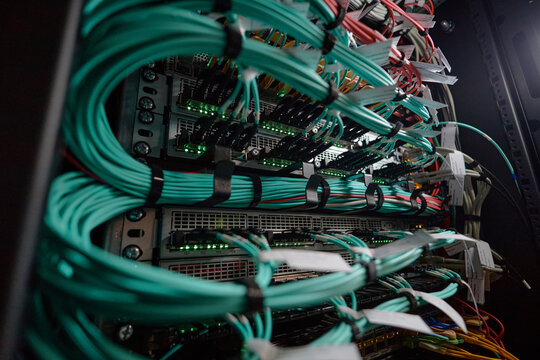 Closeup showing network cables connecting to server rack in data center, multiple teal and red wires organized and plugged into hardware, green indicator lights illuminated on equipment