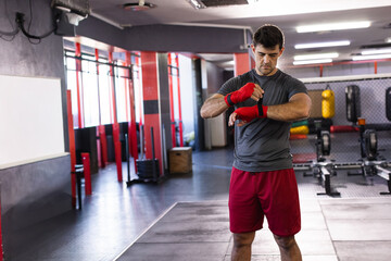 Mid adult man wrapping red hand wraps around wrists at gym with heavy bags and whiteboard © wavebreak3