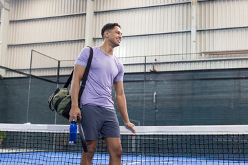 Asian man in athletic clothing leaning on net on hardcourt holding racket, bottle, carrying duffel © wavebreak3