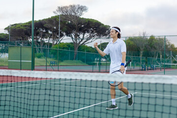 Fototapeta premium Asian man celebrating by net on green tennis court wearing white, holding racket, copy space