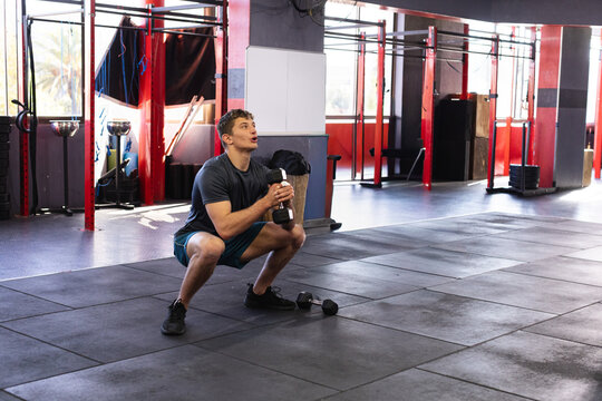 Male wearing navy shirt performing goblet squat holding dumbbell at chest on gym floor, copy space