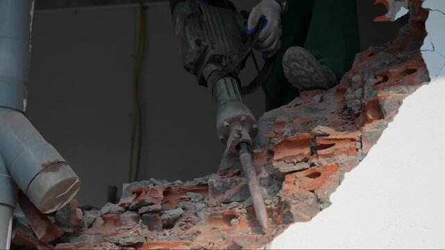 Construction and renovation. Person builder works with electrical jackhammer equipment and demolishing brick wall in building, male worker using hammer drill tool to destroy wall in old house.