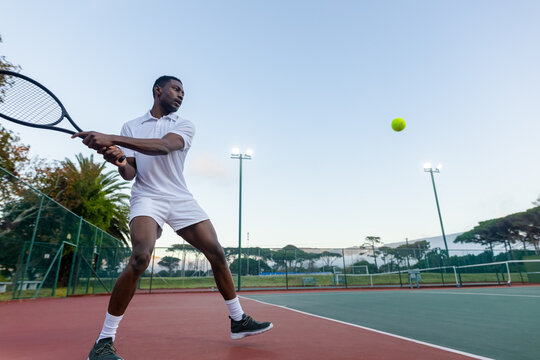 African-American male preparing two-handed backhand on court in white holding racket at yellow ball