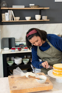 Mature baker wearing olive-apron spreading icing on layered cake at kitchen counter with spatula