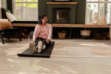 Fototapeta premium Middle-aged Asian woman sitting on mat in living room near fireplace, wearing pink hoodie, leggings