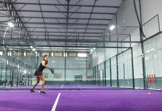 Adult Indian woman in tanktop swinging padel racket, hitting yellow ball on purple turf, copy space
