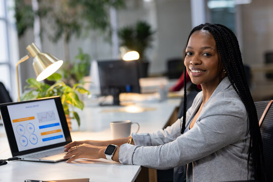African American woman in gray blazer analyzing laptop graphs on office desk with lamp, copy space