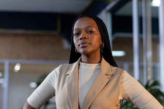 African American woman standing with braided hair and hoop earrings wearing beige vest in office