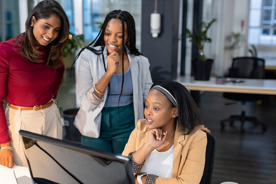 Diverse female colleagues collaborating around computer monitor in open office with potted plants