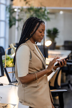 African American woman leaning against desk checking smartphone in modern office with laptop
