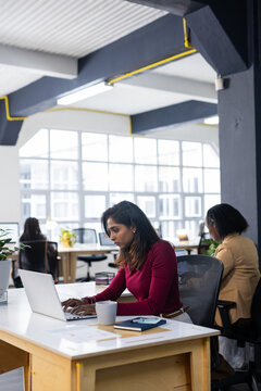 Indian woman typing on silver laptop at open-plan office desk with coffee mug and notebook