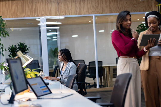 Diverse female colleagues typing on keyboards and pointing at tablet on communal desk in workspace