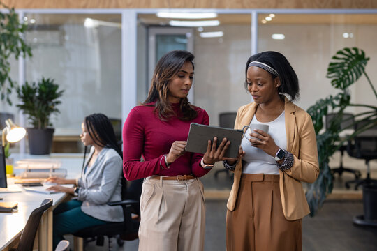 Diverse female coworkers reviewing tablet at modern open-plan office with coffee mug, desk lamp