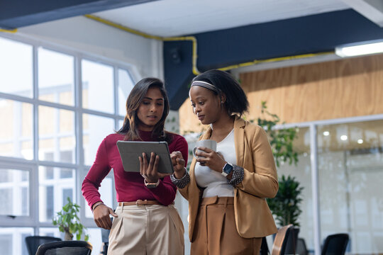 Diverse female colleagues reviewing content on tablet standing in open office with white coffee mug