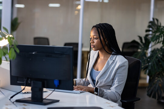 African American woman wearing grey blazer typing at office desk with monitor keyboard desk lamp