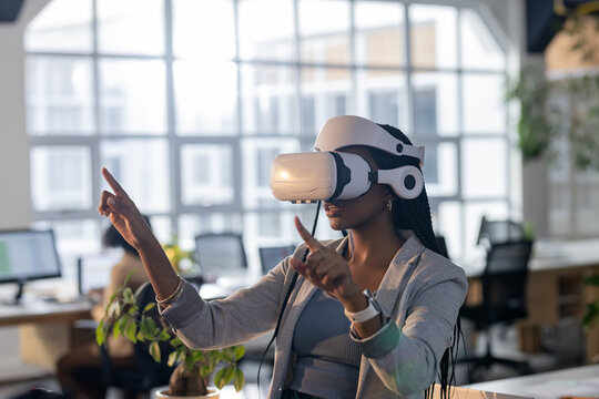 Woman wearing VR headset gesturing at desk with monitors and potted plant in open-plan office