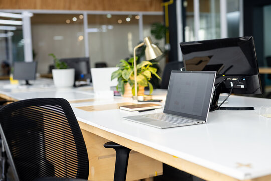 Silver laptop sitting next to black monitor on white desk with plant in open-plan office