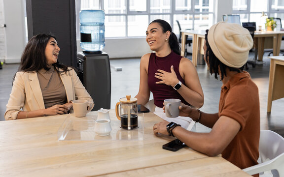 Diverse coworkers chatting around wooden table in modern office break area with French press coffee