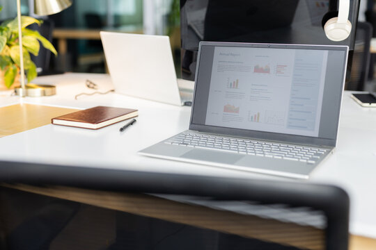 Silver laptop is displaying annual report on desk with notebook, pen, plant, headphones and lamp
