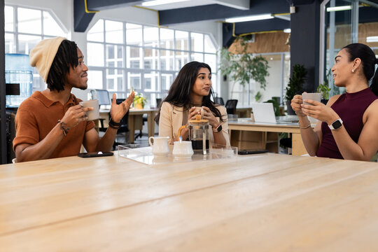 Diverse coworkers drinking French press coffee and chatting around table in modern break area