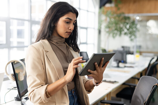 Asian woman standing at open-plan office desk holding tablet near monitor and headphones