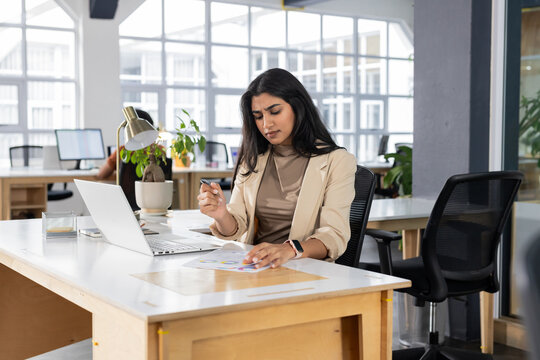 Indian businesswoman holding pen and writing on printed charts at modern office desk with laptop