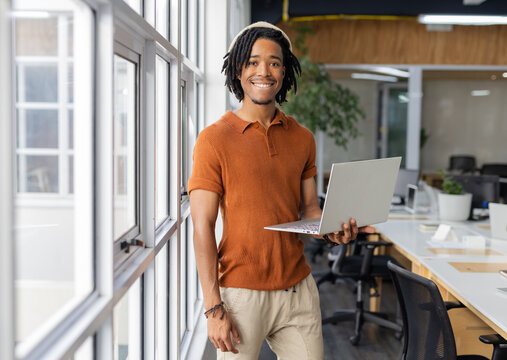 African American man leaning on windows and holding silver laptop near desks in open-plan office