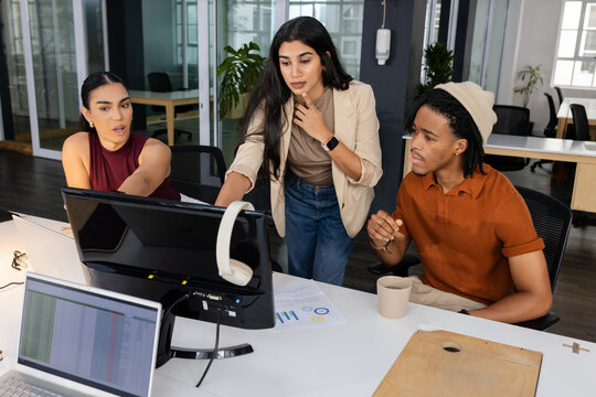 Diverse coworkers collaborating around desk in office with monitors, charts and coffee mug