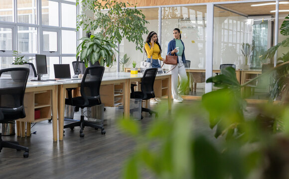 Diverse female coworkers walking past wooden desks in open office holding smartphone and coffee cup