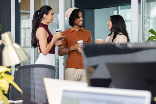 Diverse coworkers chatting and holding disposable coffee cups in open office near computer monitors
