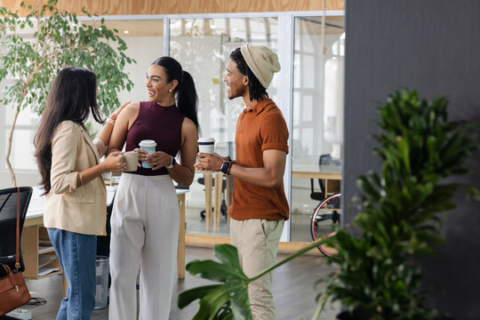 Diverse coworkers standing in office break area holding ceramic mug and takeaway cups chatting