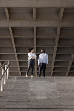 Diverse female friends in activewear standing atop concrete stairway under coffered ceiling