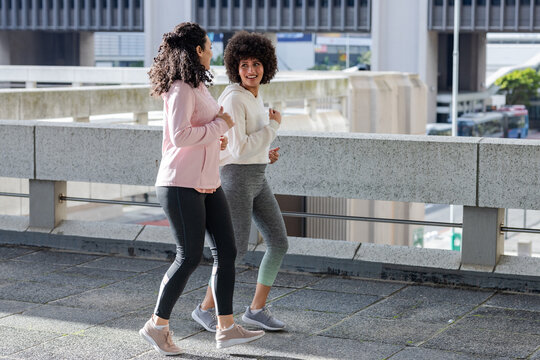 Diverse female friends walking briskly, chatting on walkway by concrete barrier wearing sneakers