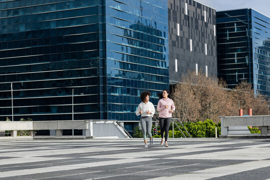 Diverse female friends jogging in sportswear on plaza, passing concrete bench, metal railing