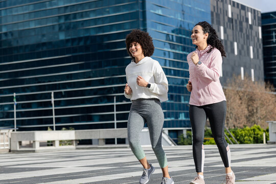Diverse female friends running along striped path by glass office building