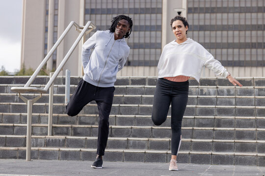 Diverse friends stretching quads while holding ankles on outdoor concrete steps, metal handrail