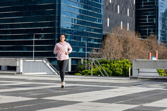 Female jogger running across urban plaza with tiles, steel railings, planter with shrubs