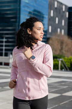 Woman with curly hair jogging across urban plaza wearing quilted jacket, checking smartwatch